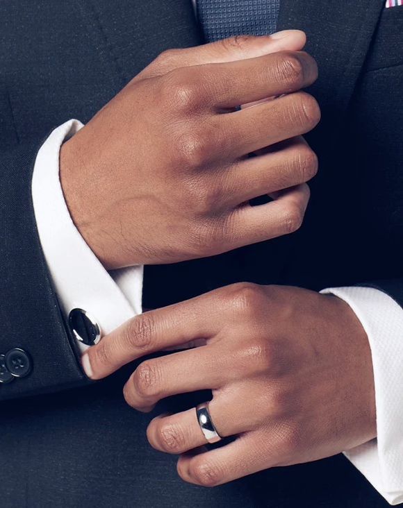 A man in a dark tailored suit and white French-cuff shirt with onyx cufflinks, wearing a polished men's wedding band