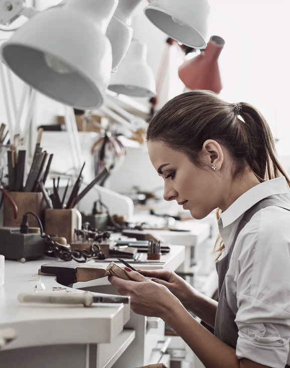 A jeweller at her workbench under industrial task lamps, focused on a piece in progress