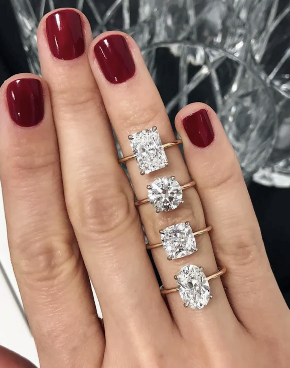 Four diamond solitaire engagement rings — radiant, round, cushion and oval — balanced on fingers with deep red lacquered nails